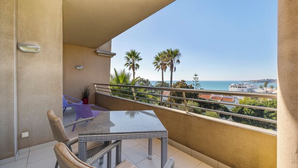 Ein Balkon mit Glastisch und Stühlen, der Blick auf die Küste mit Palmen und Gebäuden freigibt.