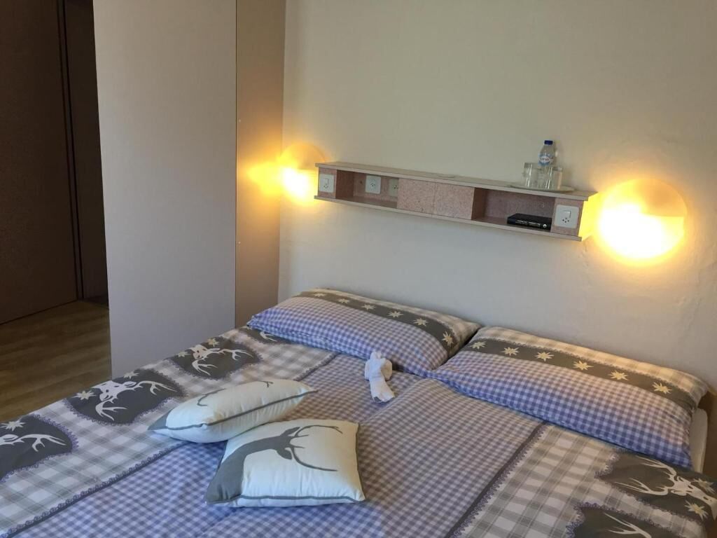 A hotel room with a bed featuring a checkered bedspread and matching pillows, a bedside table with a bottle and glass, and a wall-mounted shelf above the bed.