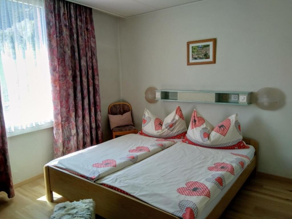 Un dormitorio con cama, una silla, una estantería con un cuadro y una ventana con cortinas.