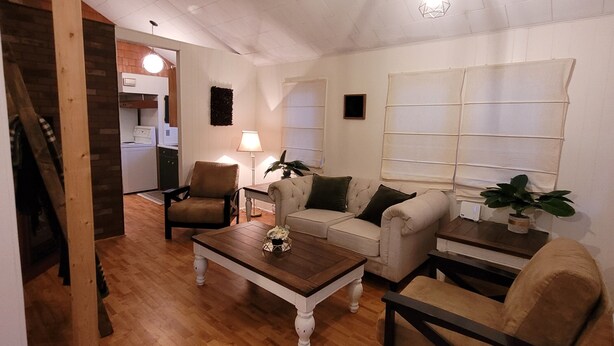 Una sala de estar acogedora con piso de madera, un sofá, un sillón y una mesa de centro. Se ve una escalera que sube a un nivel superior.