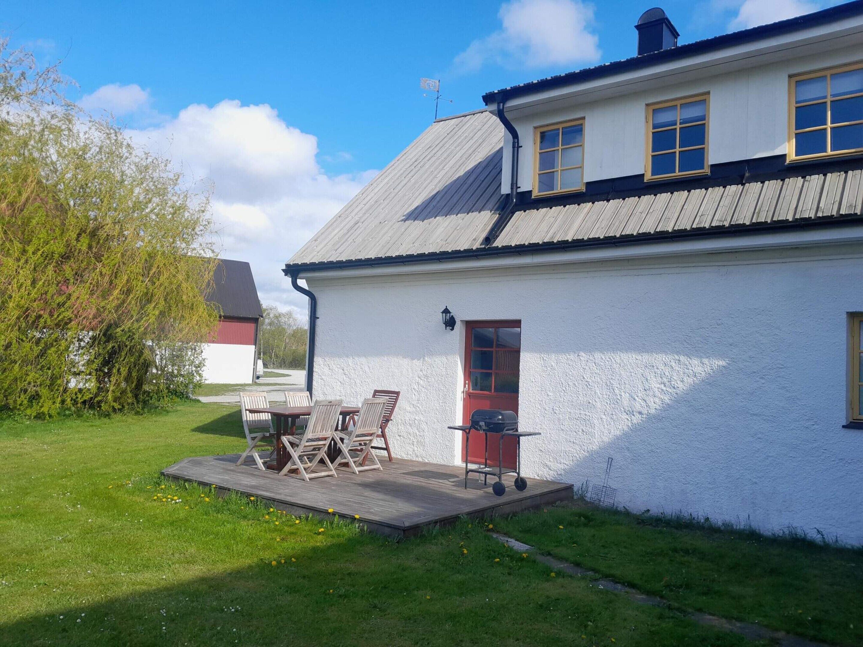 A white house with a red door, a small deck with chairs, and a grill.