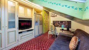 Ein Zimmer mit Fernseher, der 'NETFLIX' zeigt, einem gemusterten Teppich, einem braunen Sofa und einer Wand mit floralen Verzierungen in Grün.