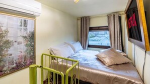 Ein kleines Zimmer mit einem Einzelbett, einem Fenster, einer grünen Geländer und einer Deckenleuchte.
