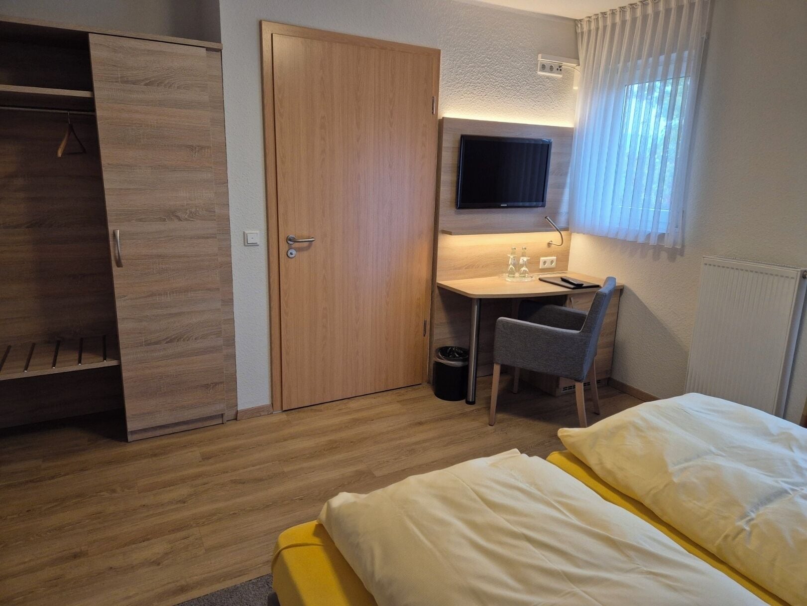 Una camera d'albergo compatta con un'armadietto in legno, una scrivania con una sedia, un letto con biancheria gialla e bianca e una TV a parete.