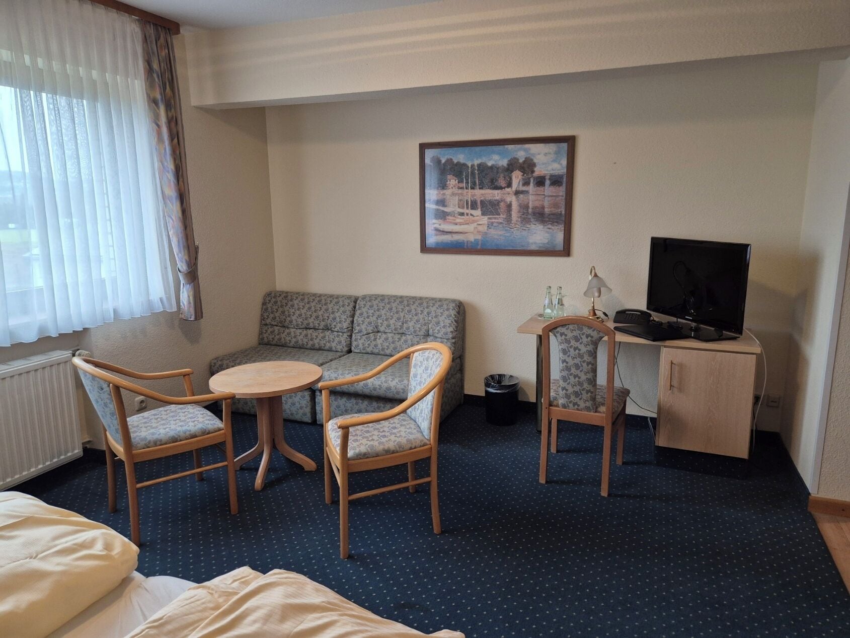A hotel room with a bed, a sofa, two chairs, a small table, a desk with a computer and lamp, a painting on the wall, and a window with curtains.