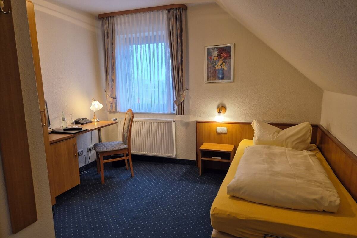 A hotel room with a bed, a desk with a chair, a radiator, a window with curtains, and a painting on the wall.