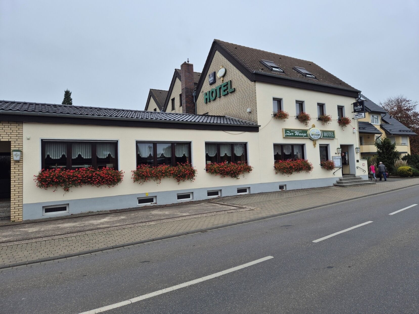 A two-story building with a sign indicating it is a hotel. There are flower boxes on the windows, and a restaurant sign is visible on the side.