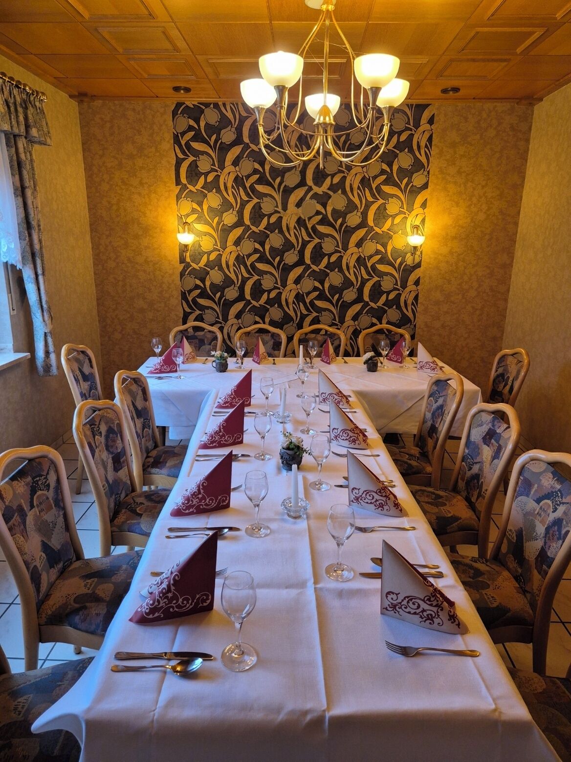 Une salle à manger avec une longue table dressée pour un repas, entourée de chaises rembourrées, d’un lustre et d’un papier peint à motifs.