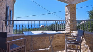 Un balcón con pared de piedra, una mesa y dos sillas. Una lámpara está sobre la mesa. El balcón tiene vista a un cielo azul despejado y a una costa lejana.