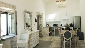 Amplia sala de estar con una mesa de comedor, una silla de mimbre blanca, una chimenea de piedra y una cocina con armarios blancos y electrodomésticos modernos.