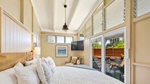 A bedroom with a large bed, a TV mounted on the wall, and a view of the outdoor area through a sliding glass door. - Oceanview Cottage | Walk to Poipu & Brennecke's Beach (Koloa)