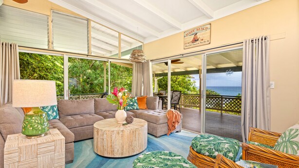 A spacious living area with a sofa, a coffee table, and a wicker chair. There is a large window with a view of the ocean. - Oceanview Cottage | Walk to Poipu & Brennecke's Beach (Koloa)