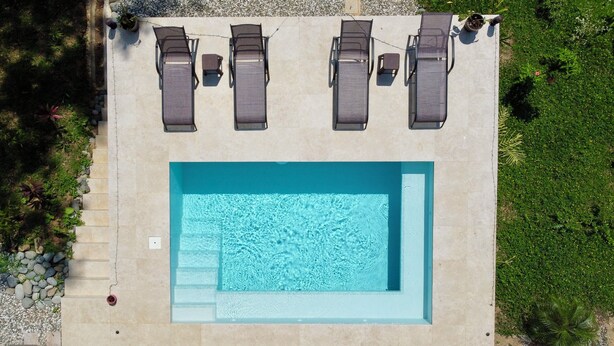 An aerial view of a rectangular swimming pool with four sun loungers and a small table, surrounded by a stone deck and greenery.