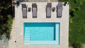 Une vue aérienne d’une piscine rectangulaire, bordée de quatre chaises longues et d’une petite table, le tout entouré d’une terrasse en pierre et de verdure.