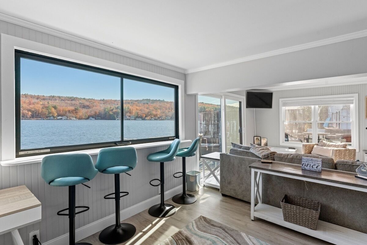 A modern living room with a large window overlooking a lake and autumn trees, a grey sofa, a coffee table, and three bar stools.