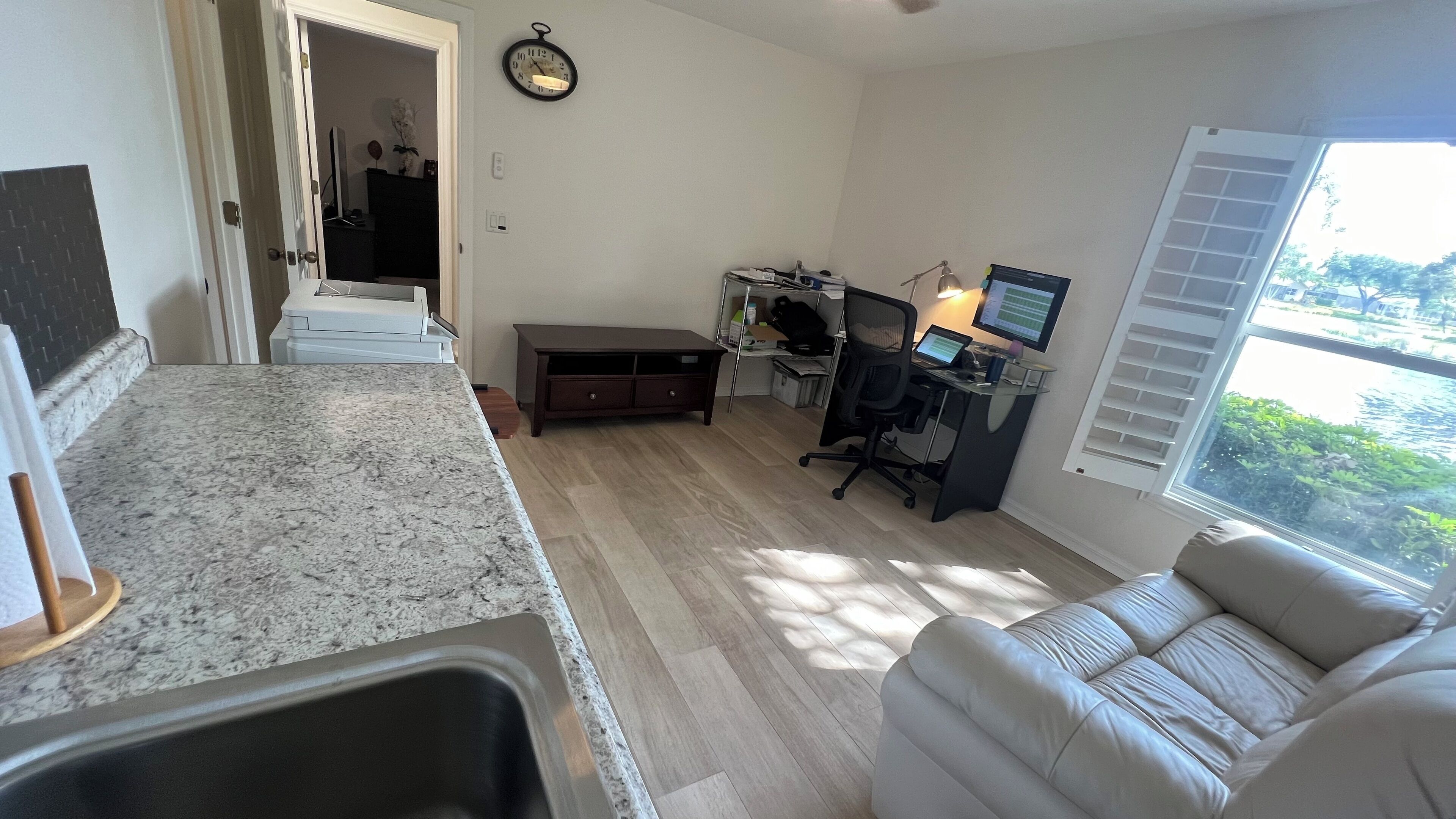 Una sala de estar con un sofá, una zona de cocina con encimera de granito, una mesa de comedor y un escritorio con monitor.
