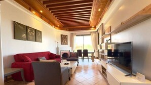 A living room with a red sofa, grey armchair, and a flat-screen TV on a white stand. There are three framed artworks on the wall and a fireplace in the background. - Magnificent T2 with swimming pool (Marrakech)