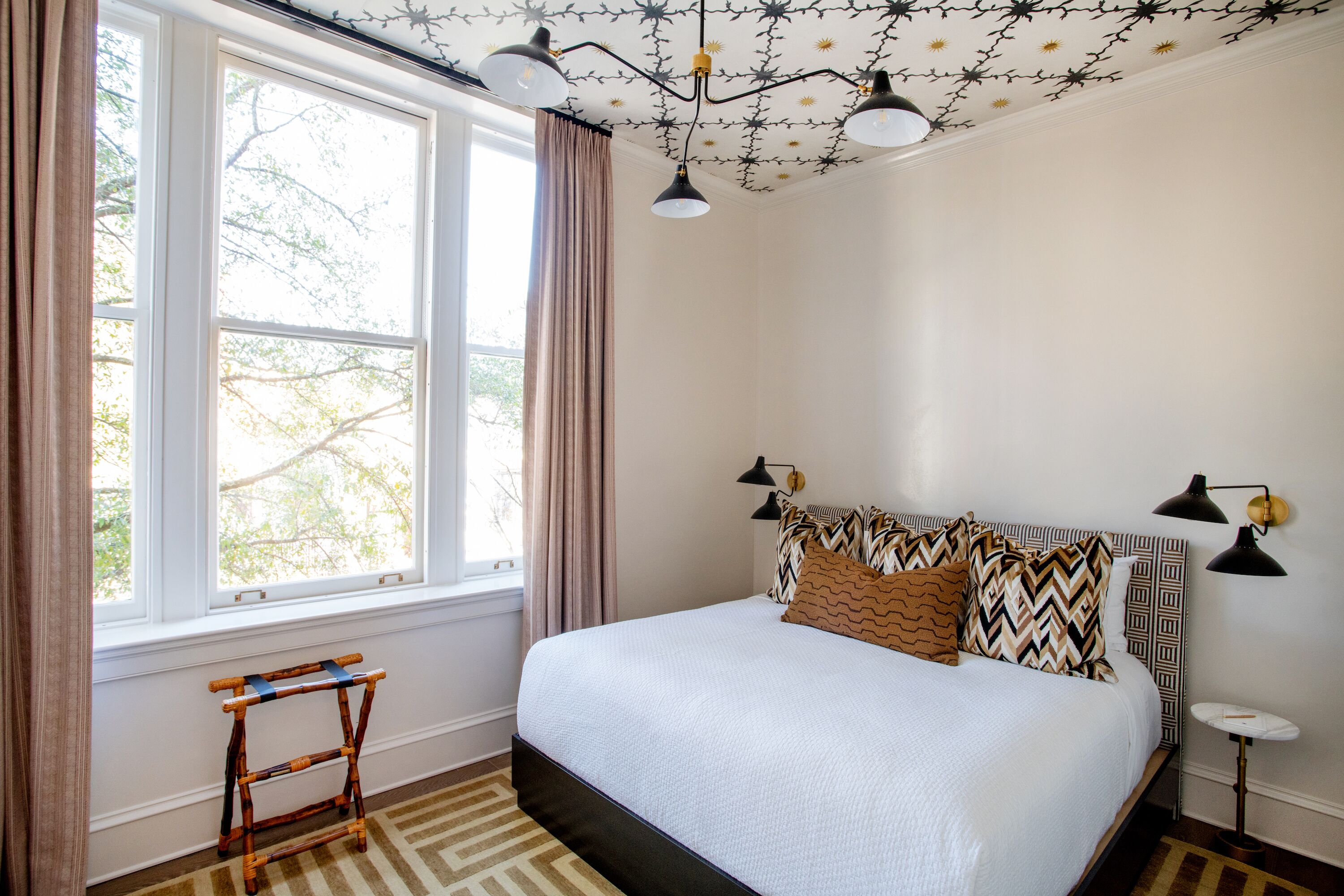A bedroom with a large bed, a window with curtains, a hanging light fixture, and a small table.