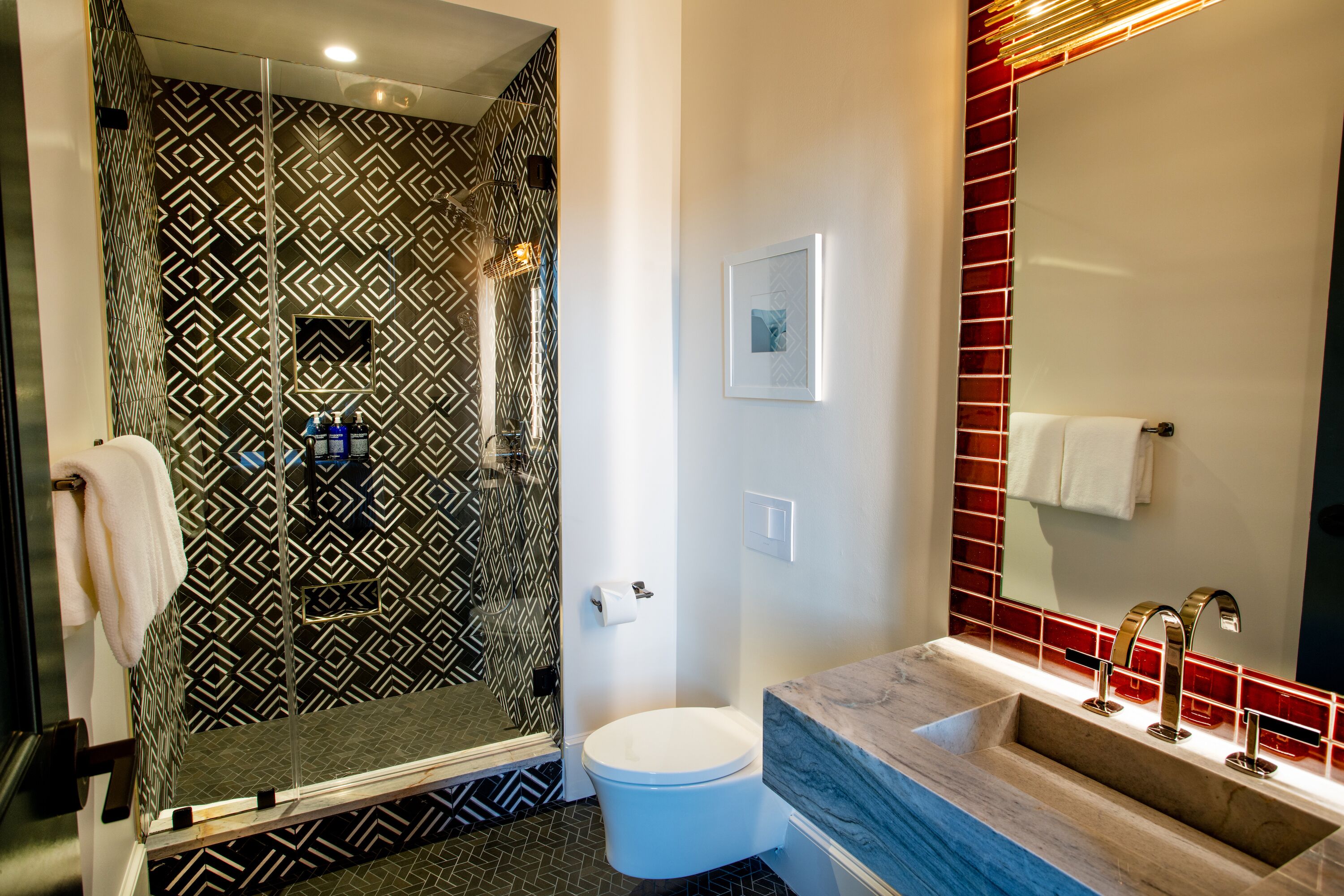 Un baño con pared de ducha estampada, inodoro, lavamanos con encimera de mármol y espejo con marco rojo.