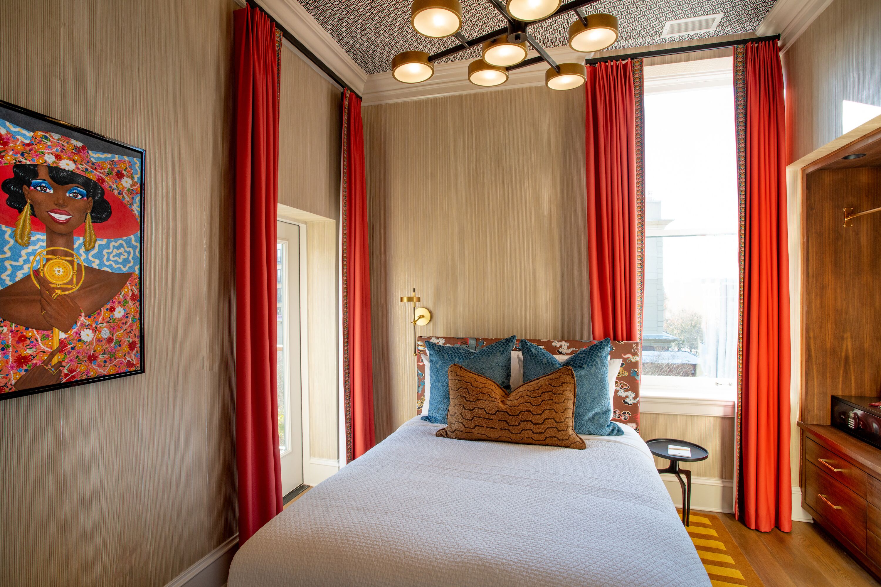 A hotel room with a large bed, red curtains, a painting on the wall, and a view of the city through the window.