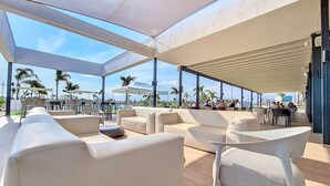 An outdoor seating area with white sofas and tables, large windows, and palm trees in the background. - Ground floor in front of the pool, south facing. (Torre Pacheco)