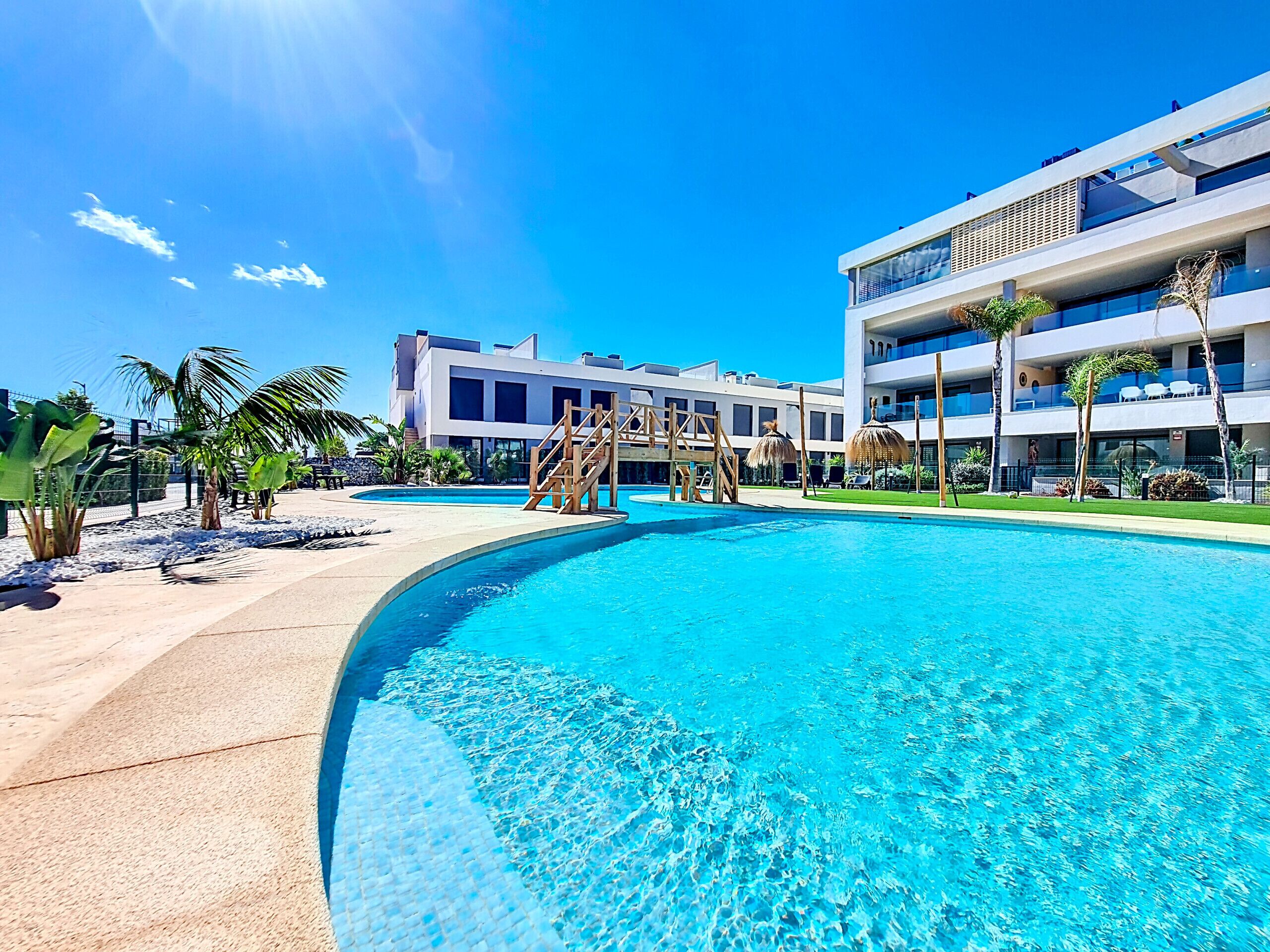 Um hotel moderno com uma piscina grande, palmeiras e um céu azul límpido.