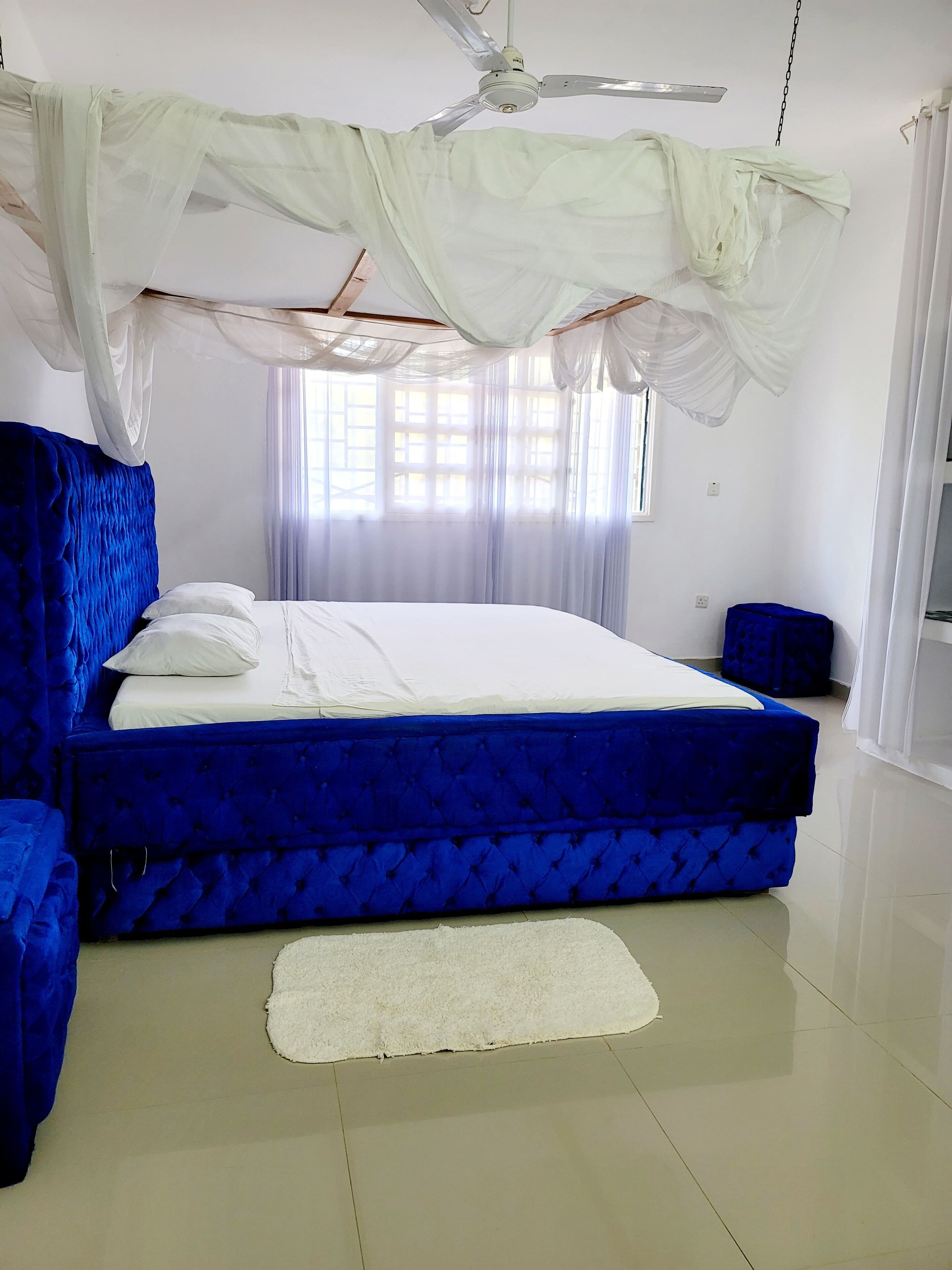 A bedroom with a large bed, a ceiling fan, sheer curtains, and a white rug.