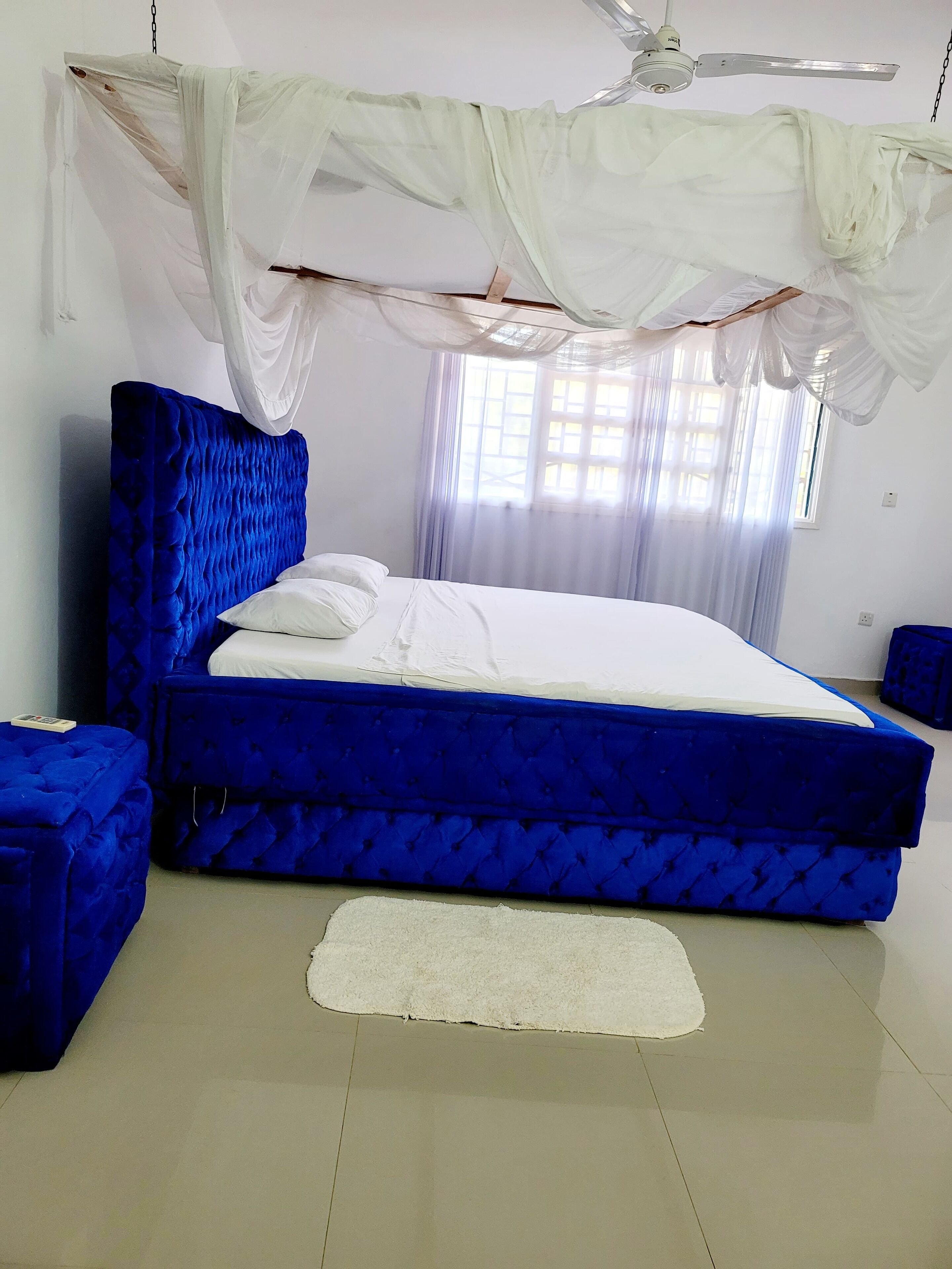 Une chambre à coucher avec un lit recouvert d’un tissu bleu, du linge de lit blanc, un tapis blanc et des rideaux légers.