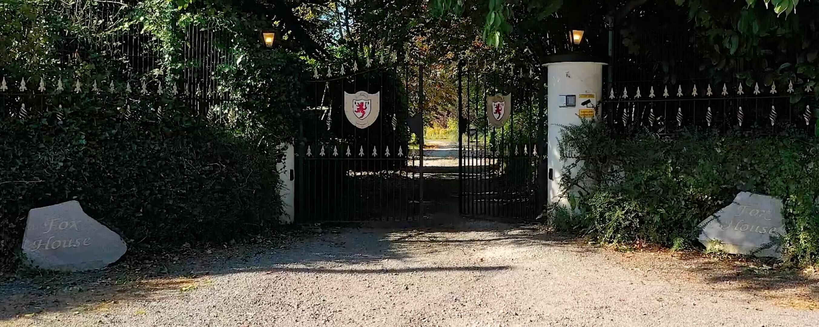 A gated entrance with a coat of arms, a stone sign reading 'Fox House,' and a gravel path leading through.