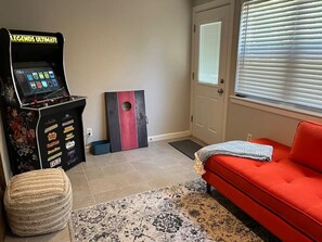 Una habitación con un sofá rojo, una consola de videojuegos, una pared con títulos de juegos, una puerta y una ventana con persianas.