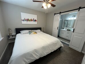 A bedroom with a bed, a ceiling fan, a nightstand, a bedside table, a wall-mounted mirror, and a door leading to a bathroom. - Rt 66 Retreat - Near Fairgrounds (Tulsa)