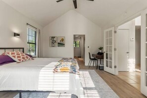 A bedroom with a large bed, a desk, and a chair, with a view of a garden through the window.