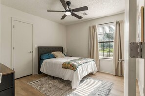 Ein Schlafzimmer mit einem Bett, einem Deckenventilator, einem Fenster mit Vorhängen und einer Tür.