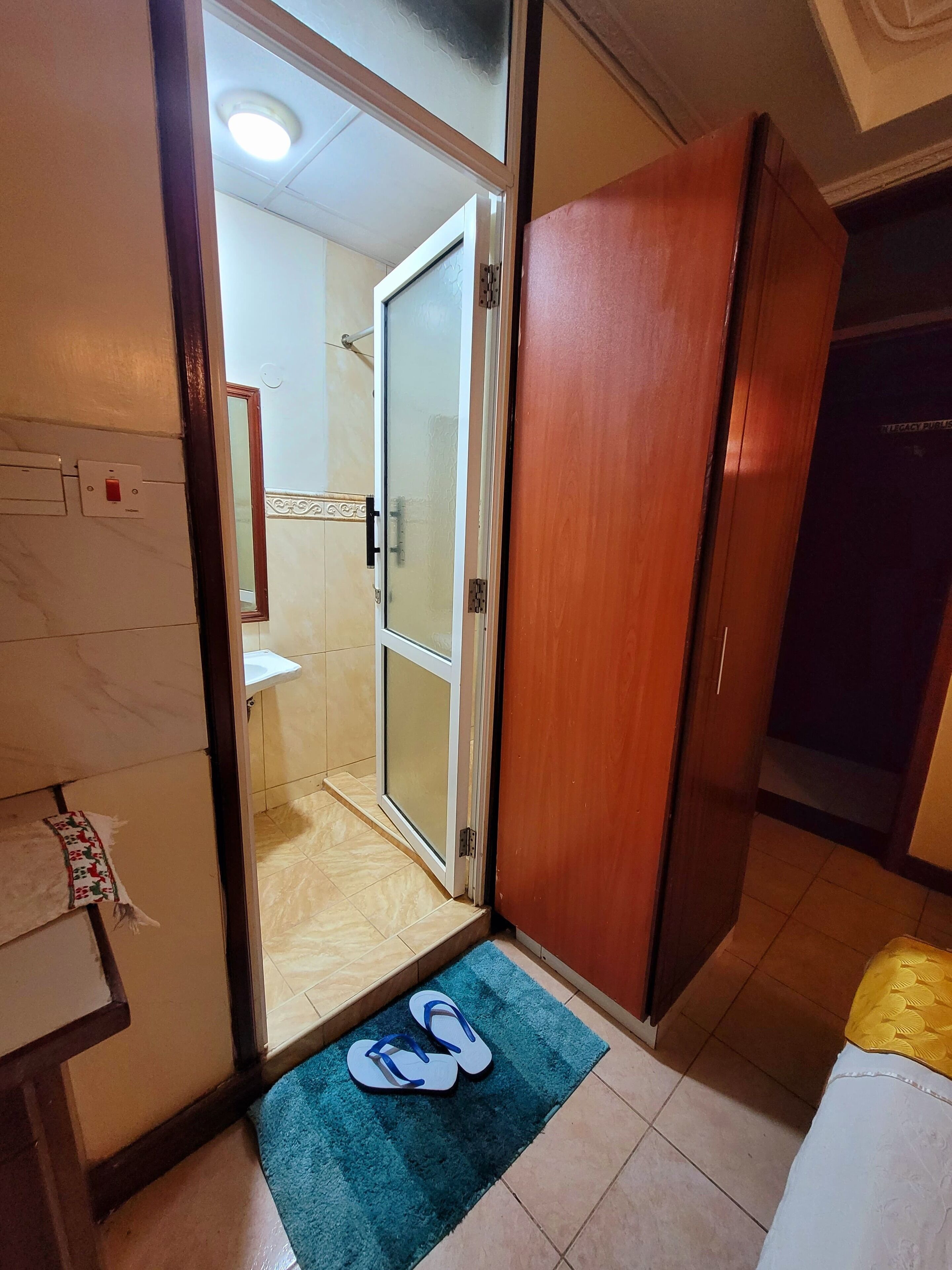 A hotel room with a wooden wardrobe, a bathroom with a glass door, and a blue mat with flip-flops on the floor.