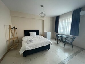 Habitación de hotel con cama, escritorio, silla, refrigerador pequeño y ventana con cortinas.