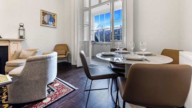 A modern living room with a fireplace, comfortable seating, a dining area with a table set for two, and a large window offering a city view. - Upscale Urban Haven: Luxury Flat (London)