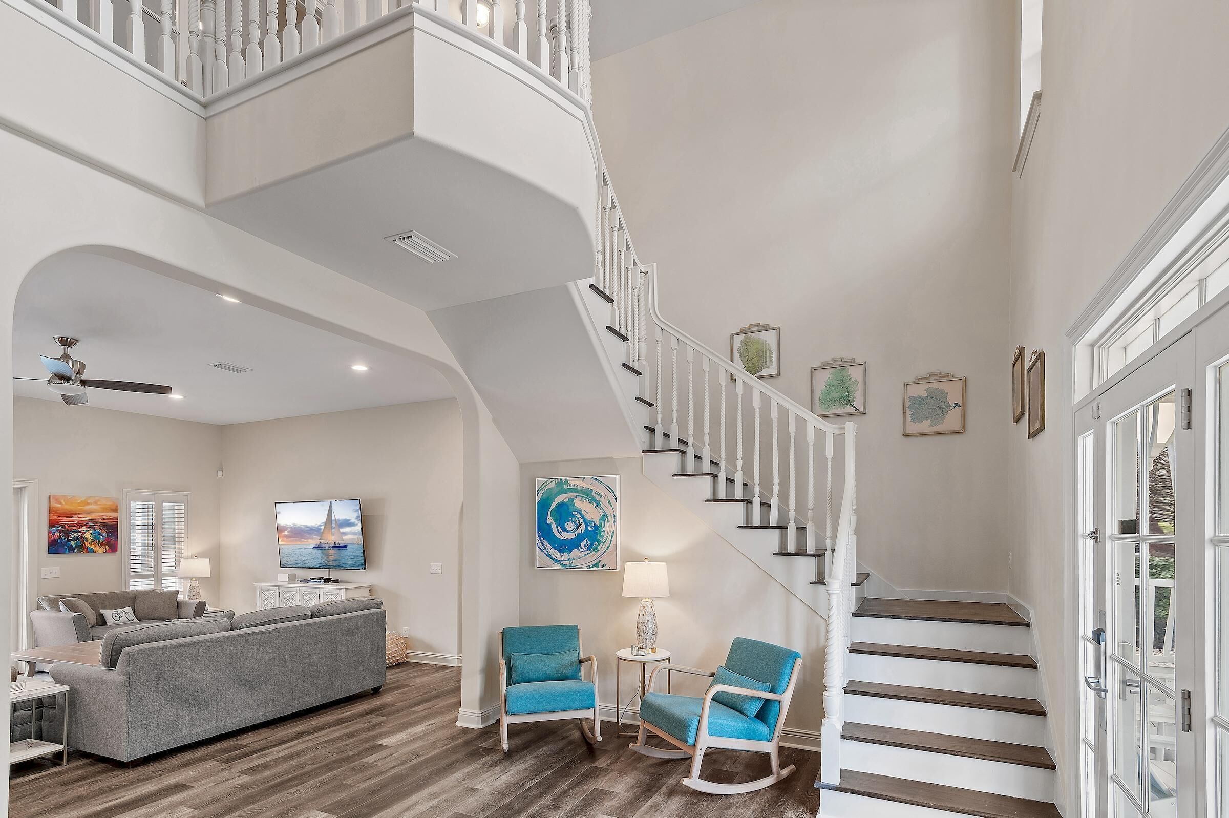 A spacious living room with a gray sofa, two rocking chairs, a TV, and artwork on the walls. A staircase with white railings leads to an upper level.