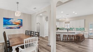A spacious dining area with a wooden table and white chairs, a kitchen with white cabinets and a green backsplash, and a large painting of a beach scene on the wall. - Sapphire by the Sea (Miramar Beach)