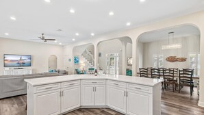 A modern kitchen with white cabinetry, a large island, and a dining area with wooden chairs. - Sapphire by the Sea (Miramar Beach)