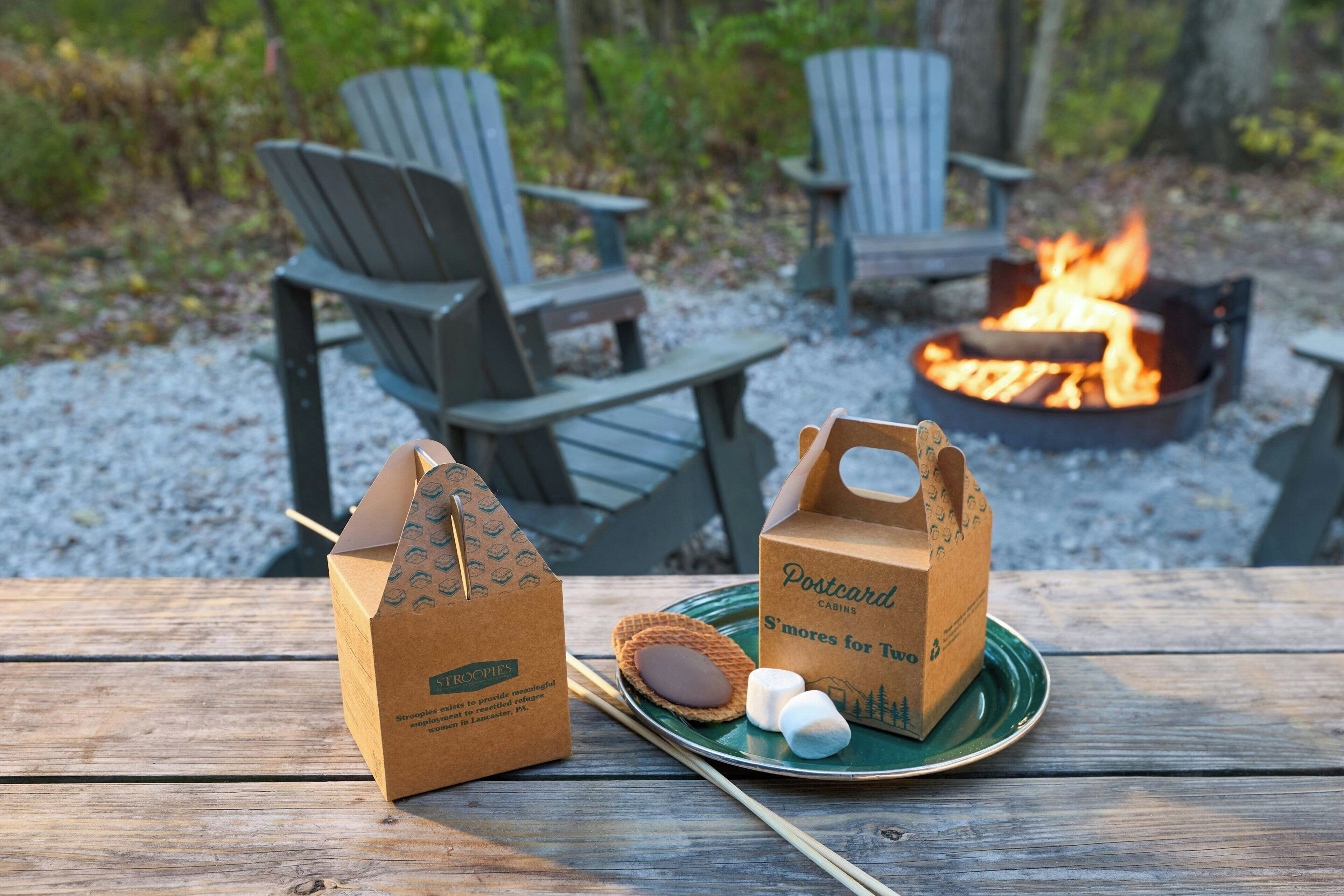 A wooden picnic table with two Adirondack chairs, a fire pit, and two paper bags labeled "Postcard Cabins" with marshmallows and cookies.