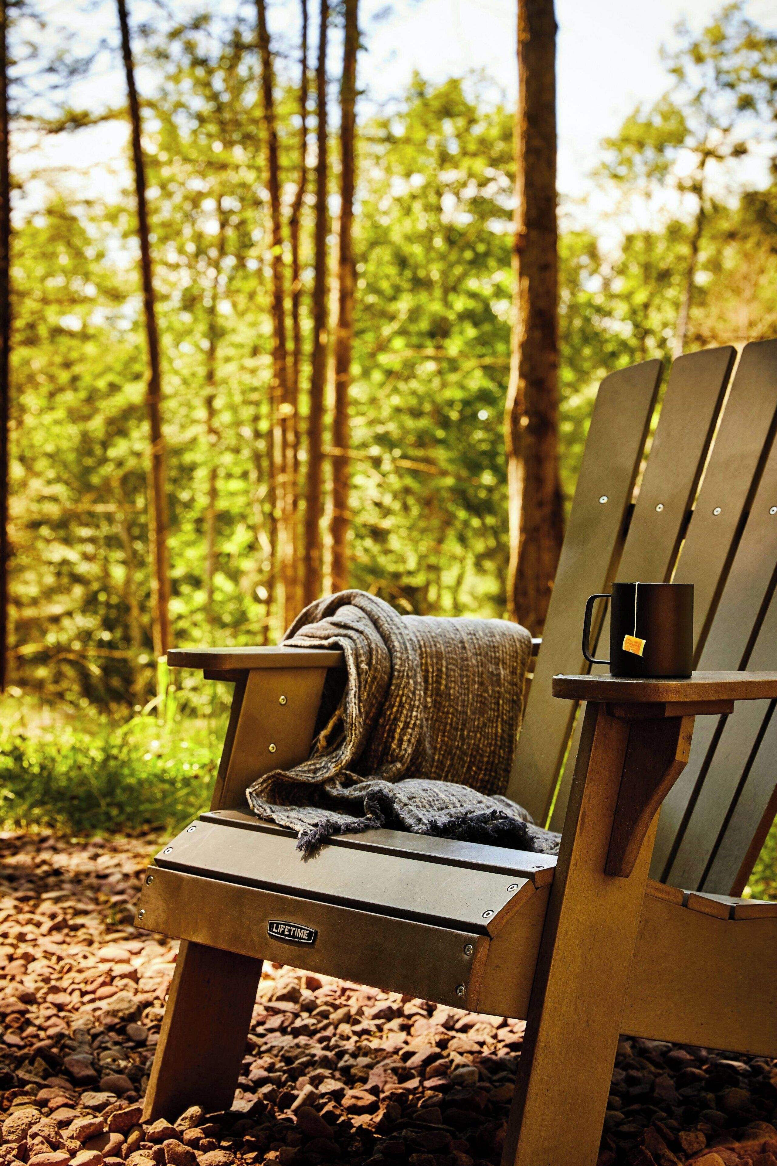 A wooden Adirondack chair with a blanket and a mug in a forest setting.