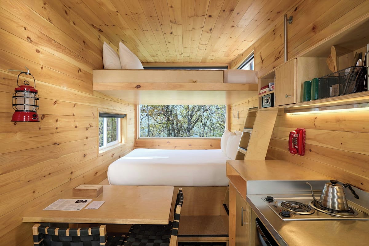 Uma cabana compacta com beliche, uma cozinhenha com fogão e uma janela com vista para árvores.