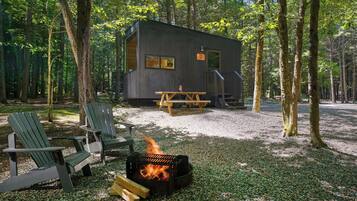 A cabin with a fire pit, two Adirondack chairs, and a picnic table in a forest setting.
