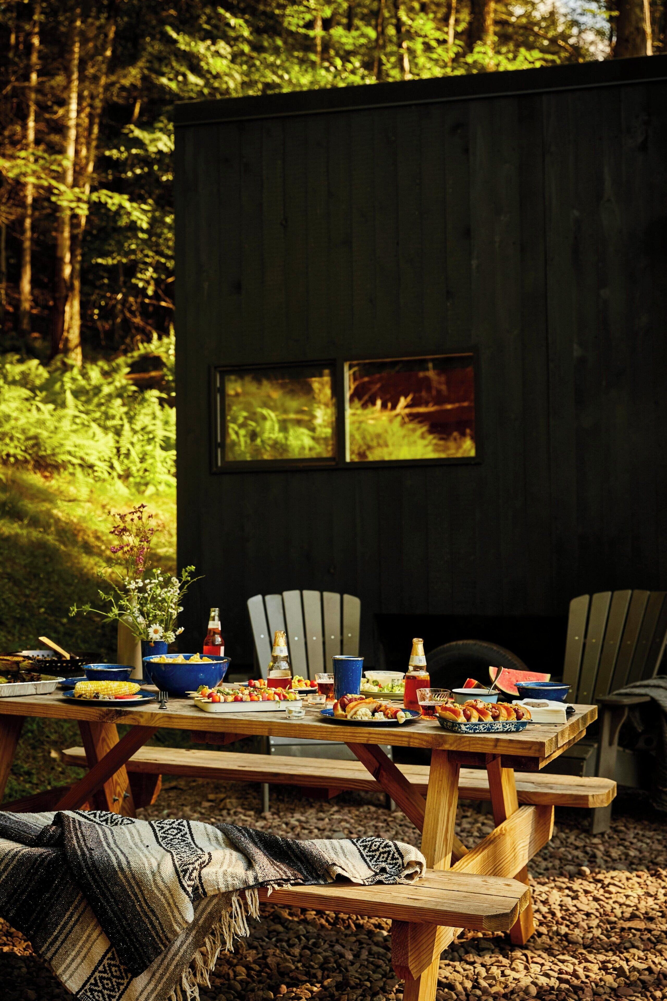 A rustic wooden cabin with a dining table set for a meal, surrounded by forest.