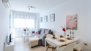 A modern living room with a grey sofa, a white dining table with chairs, and a large window with sheer curtains. - 1bed & Sofabed-seaside Apt-quiet Location-parking (Alicante)