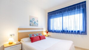 A neatly arranged hotel room with a bed, bedside table, lamp, and a window with blue curtains. - 1bed & Sofabed-seaside Apt-quiet Location-parking (Alicante)