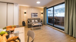 A modern living room with a wooden wall, a sofa, a coffee table, and a dining area with a basket of bread and fruit. - Lagrange Vacances Le Cristal de Piau  (Aragnouet)