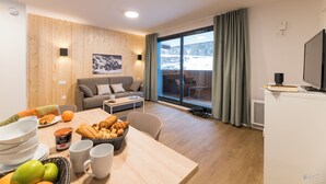 A modern living room with a wooden wall, a sofa, a dining table with breakfast items, and a view of a snowy landscape through large windows. - Lagrange Vacances Le Cristal de Piau  (Aragnouet)