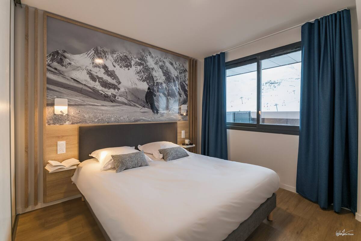 Quarto de hotel com uma grande moldura retratando uma paisagem de montanha, cama bem arrumada com almofadas, mesa de cabeceira com abajur e janela com cortinas.