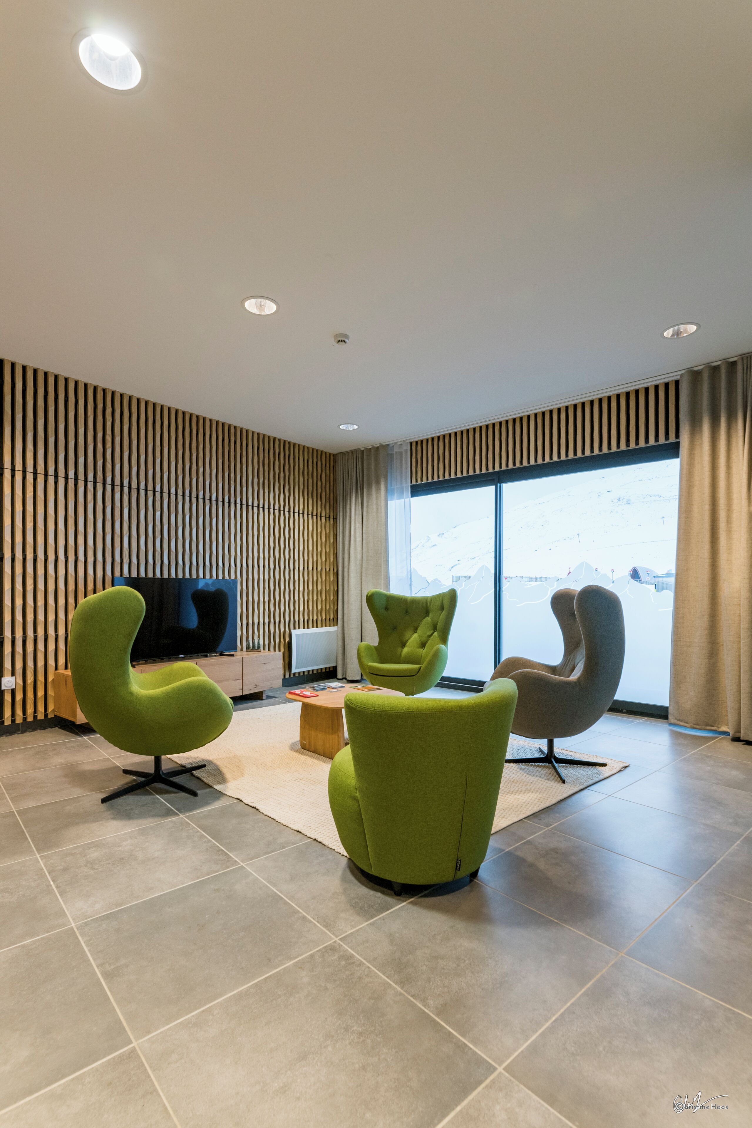 Una sala de estar moderna con un televisor de pantalla plana, dos sillones verdes, un mueble de madera y un ventanal con vistas a un paisaje nevado.
